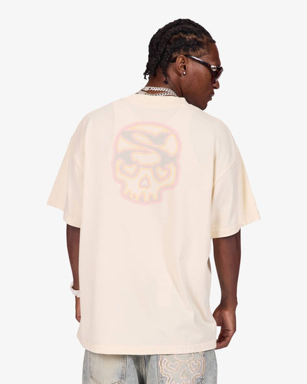 A man shown from the back wearing an oversized cream-colored t-shirt with a large, faded pink and orange skull graphic on the back, paired with light-wash jeans.