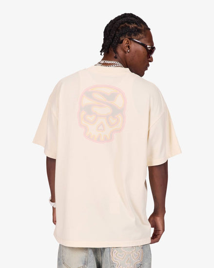 A man shown from the back wearing an oversized cream-colored t-shirt with a large, faded pink and orange skull graphic on the back, paired with light-wash jeans.