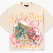 A cream-colored short-sleeve graphic t-shirt featuring a large front print of two skulls in green and pink with flames and the text 'SUGARHUL'.