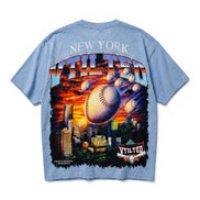 A light blue short-sleeve t-shirt shown from the back, featuring a large graphic print of a baseball flying over a New York City skyline at sunset, with 'NEW YORK' and 'V Tilted' text.