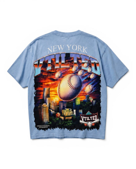 A light blue short-sleeve t-shirt shown from the back, featuring a large graphic print of a baseball flying over a New York City skyline at sunset, with 'NEW YORK' and 'V Tilted' text.