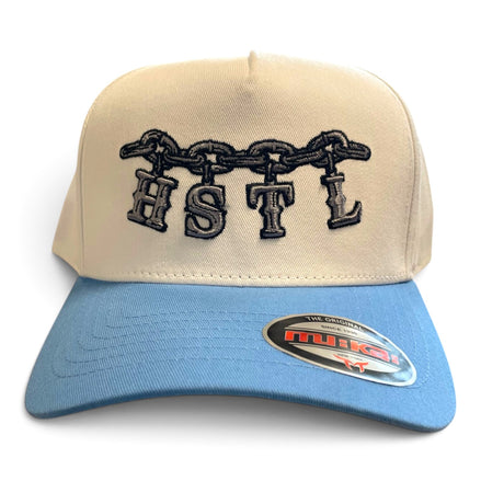 A structured trucker-style baseball cap with a cream-colored crown and a light blue brim. The front crown features black embroidered lettering spelling 'HSTL' beneath a black chain motif.