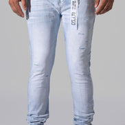 Light wash blue distressed skinny jeans with a vertical white brand tag on the left thigh, modeled with black sneakers and a black t-shirt against a gray studio background.