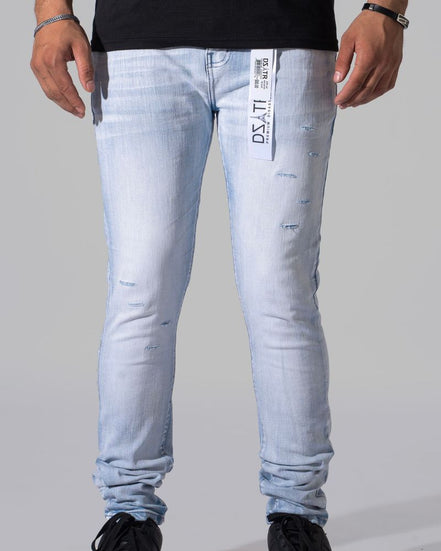 Light wash blue distressed skinny jeans with a vertical white brand tag on the left thigh, modeled with black sneakers and a black t-shirt against a gray studio background.