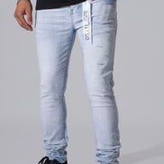 Light wash blue slim-fit jeans with distressed details and a vertical brand tag on the front pocket, shown on a model wearing a black t-shirt and black sneakers against a gray studio background.