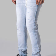 Light wash blue slim-fit jeans with distressed details, worn by a model with black sneakers against a gray studio background.