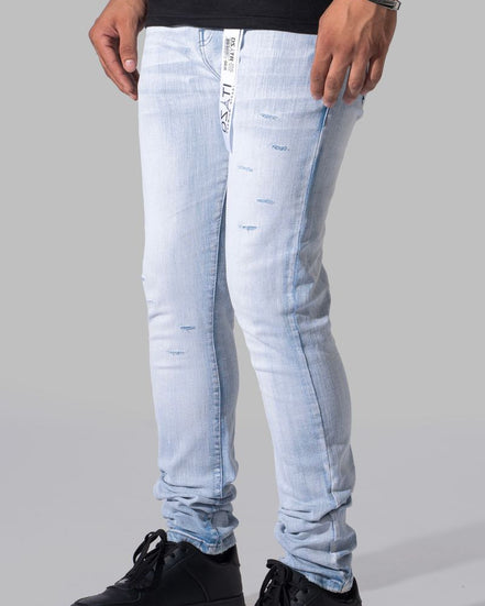 Light wash blue slim-fit jeans with distressed details, worn by a model with black sneakers against a gray studio background.