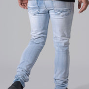 Rear view of a person wearing light-wash slim-fit jeans, a black t-shirt, and black shoes against a plain gray studio background.