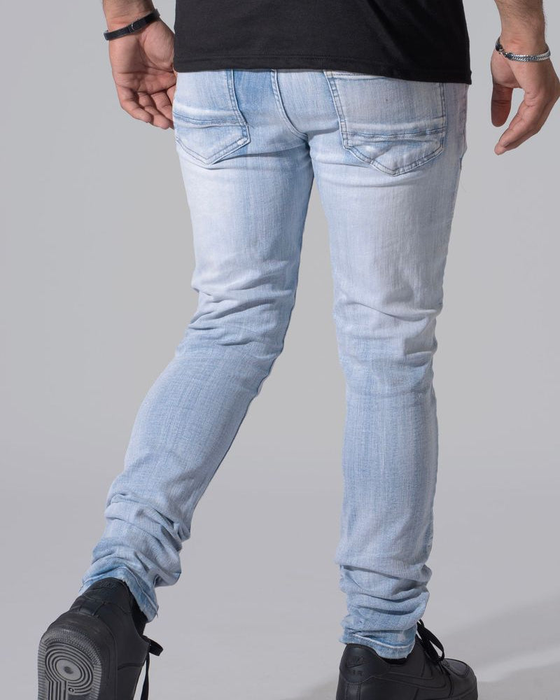 Rear view of a person wearing light-wash slim-fit jeans, a black t-shirt, and black shoes against a plain gray studio background.