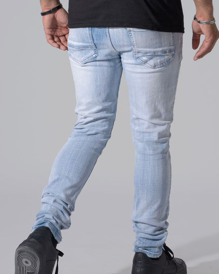 Rear view of a person wearing light-wash slim-fit jeans, a black t-shirt, and black shoes against a plain gray studio background.