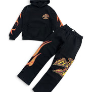 A two-piece kids' tracksuit set consisting of a black hoodie and matching sweatpants, both decorated with vibrant orange and yellow flame graphics and a small chest emblem, displayed on a clean white background.
