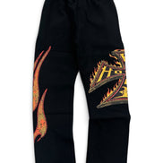 Black sweatpants featuring a large orange and yellow flame graphic on the right leg and a stylized gold and orange letter 'H' on the left leg, displayed against a plain white background.