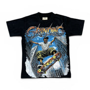 A black short-sleeve graphic t-shirt featuring a large front print of a skateboarder in mid-air between tall buildings under the word 'GRINDING' in metallic lettering.