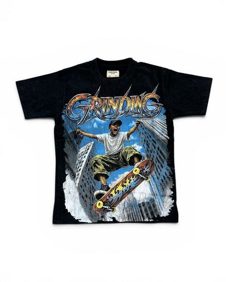 A black short-sleeve graphic t-shirt featuring a large front print of a skateboarder in mid-air between tall buildings under the word 'GRINDING' in metallic lettering.