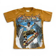 A vintage-style graphic t-shirt in a washed mustard yellow color, featuring a large front print of a skateboarder in a city with the word 'GRINDING' in stylized metallic lettering.