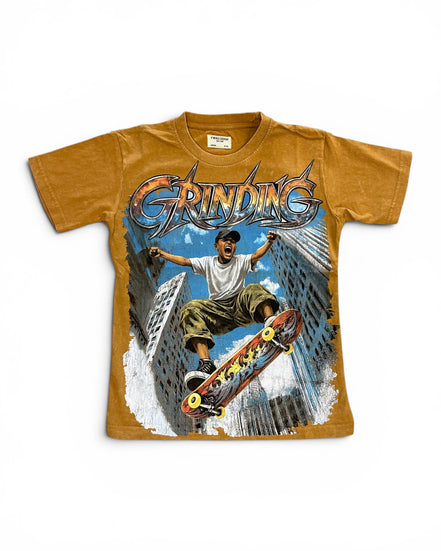 A vintage-style graphic t-shirt in a washed mustard yellow color, featuring a large front print of a skateboarder in a city with the word 'GRINDING' in stylized metallic lettering.