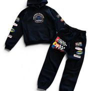 A two-piece kids' racing team hoodie and sweatpants set in dark navy blue. The hoodie features a chest graphic and various sponsor patches on the sleeves.