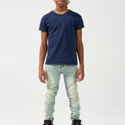 A child model wearing a navy blue crewneck t-shirt, light-wash distressed jeans, and black sneakers with red accents, standing against a plain white studio background.