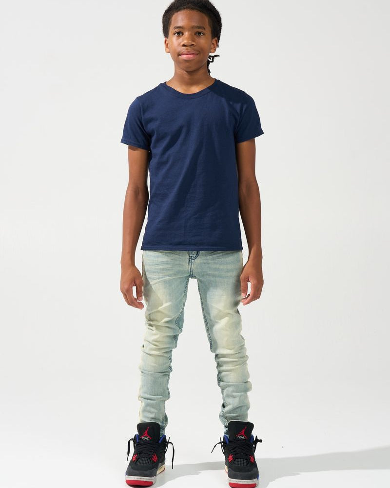 A child model wearing a navy blue crewneck t-shirt, light-wash distressed jeans, and black sneakers with red accents, standing against a plain white studio background.