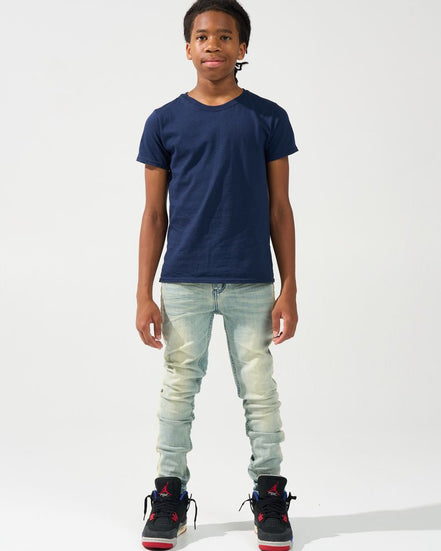 A child model wearing a navy blue crewneck t-shirt, light-wash distressed jeans, and black sneakers with red accents, standing against a plain white studio background.