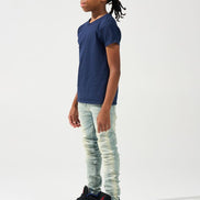 A child model shown in profile wearing a navy blue t-shirt, light-wash distressed jeans, and black sneakers with red and blue accents, set against a plain white studio background.