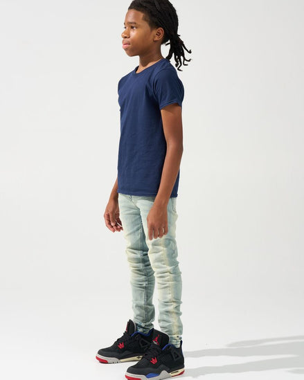 A child model shown in profile wearing a navy blue t-shirt, light-wash distressed jeans, and black sneakers with red and blue accents, set against a plain white studio background.