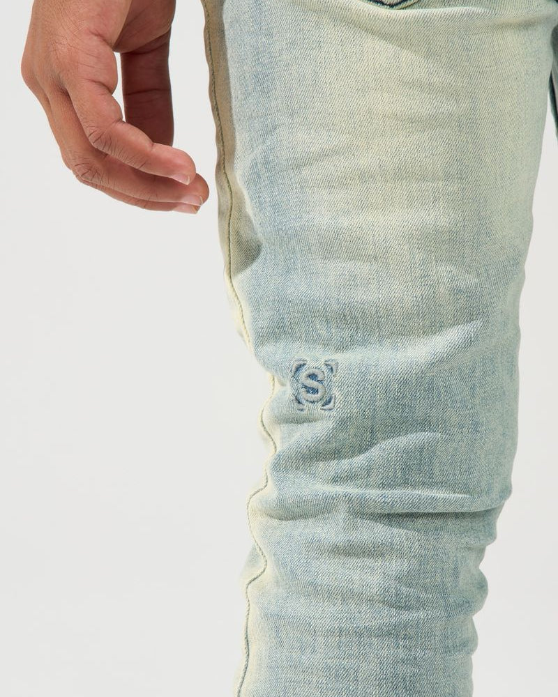 Close-up of a person's hand and thigh wearing light-wash blue denim jeans with a small embroidered logo on the upper leg.