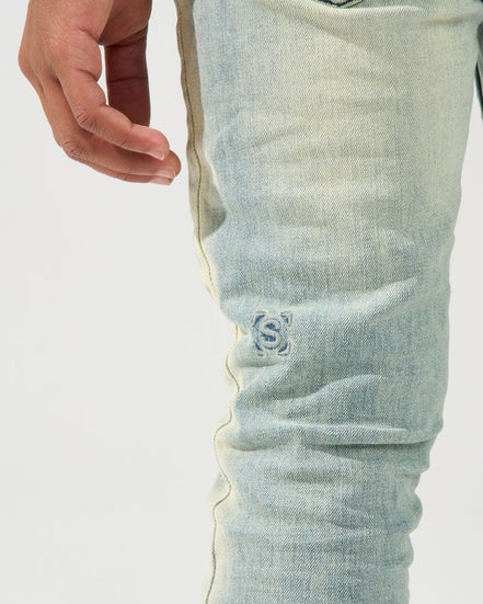 Close-up of a person's hand and thigh wearing light-wash blue denim jeans with a small embroidered logo on the upper leg.