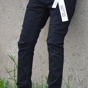 Person wearing black pants with a visible brand tag, standing on grass against a concrete wall.