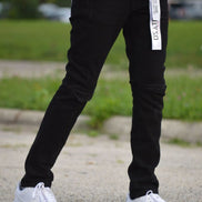 Person wearing black pants with a visible brand logo and white sneakers on a blurred outdoor background