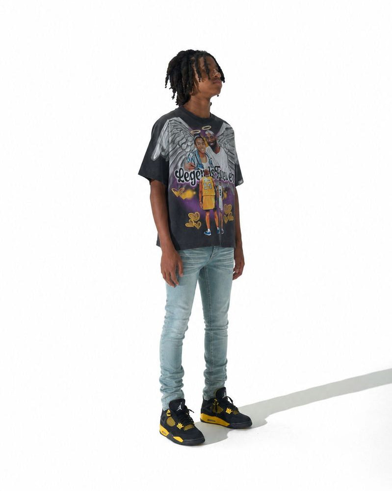 A person standing against a white studio background wearing a black graphic t-shirt, light blue jeans, and black sneakers with yellow accents.