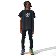 A person stands against a white studio background wearing an oversized black graphic T-shirt, dark pants, and colorful sneakers. The shirt has a white square cross-style logo on the chest.