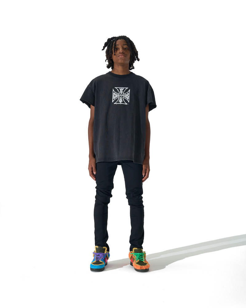 A person stands against a white studio background wearing an oversized black graphic T-shirt, dark pants, and colorful sneakers. The shirt has a white square cross-style logo on the chest.