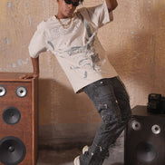 Person wearing a graphic t-shirt and dark jeans posing next to audio speakers against a textured wall.