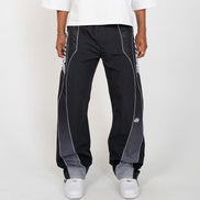 A person wearing black wide-leg track pants with gray side panels and white piping, paired with a white top and white sneakers, shown against a light gray studio background.