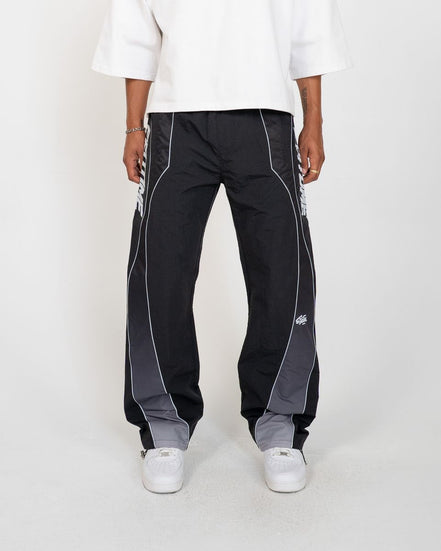A person wearing black wide-leg track pants with gray side panels and white piping, paired with a white top and white sneakers, shown against a light gray studio background.