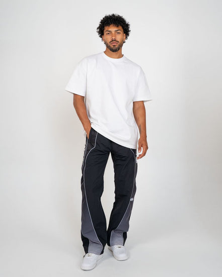A man modeling a white short-sleeve t-shirt and dark gray side-panel track pants with white piping, paired with white sneakers, against a light gray studio background.