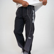 A person wearing black athletic pants with white side piping and a large white vertical 'EPITOME' logo on one leg, paired with a white t-shirt and white sneakers against a light gray studio background.