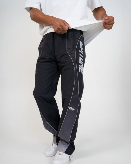 A person wearing black athletic pants with white side piping and a large white vertical 'EPITOME' logo on one leg, paired with a white t-shirt and white sneakers against a light gray studio background.