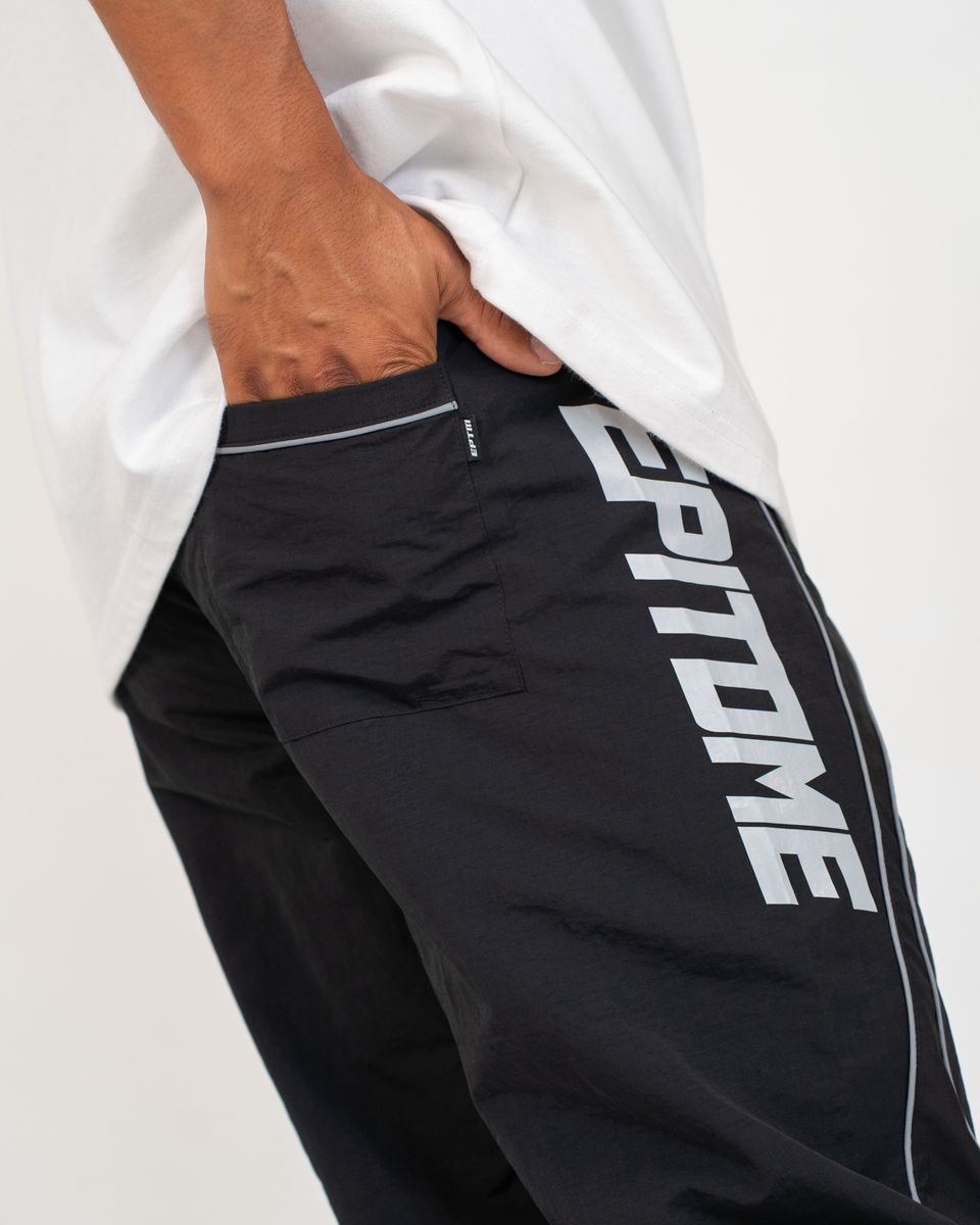 Close-up of a person wearing black athletic pants with large white 'EPITOME' lettering down the side and a white t-shirt, with one hand in the pocket.