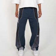 A person wearing navy track pants with dark gray side panels and white piping, paired with a white t-shirt and white sneakers, shown from the waist down against a light gray studio background.