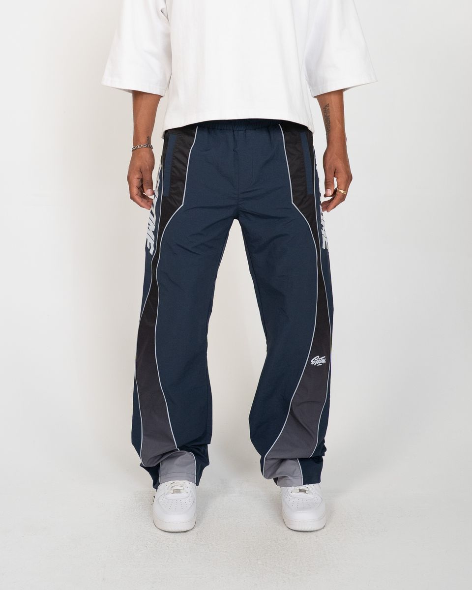 A person wearing navy track pants with dark gray side panels and white piping, paired with a white t-shirt and white sneakers, shown from the waist down against a light gray studio background.