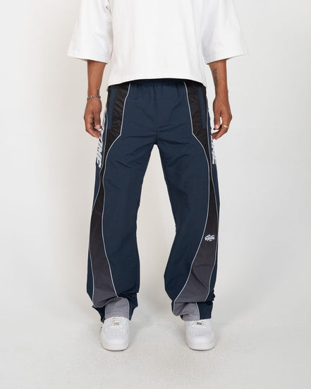 A person wearing navy track pants with dark gray side panels and white piping, paired with a white t-shirt and white sneakers, shown from the waist down against a light gray studio background.