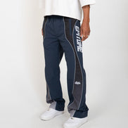 A person wearing navy and gray athletic pants with white piping and a large white vertical logo on the side, paired with a white t-shirt and white sneakers, photographed against a light gray studio background.