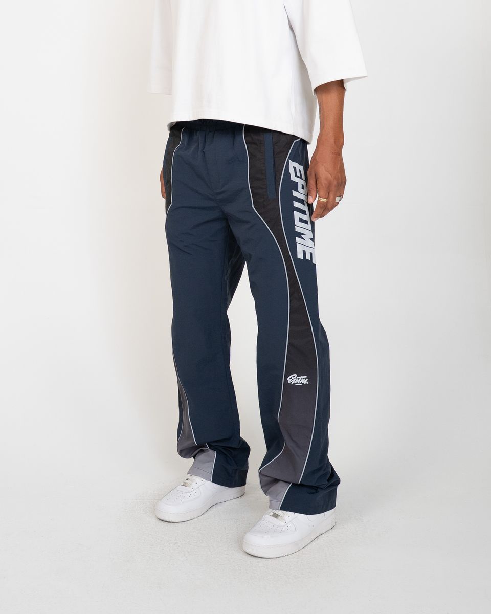 A person wearing navy and gray athletic pants with white piping and a large white vertical logo on the side, paired with a white t-shirt and white sneakers, photographed against a light gray studio background.