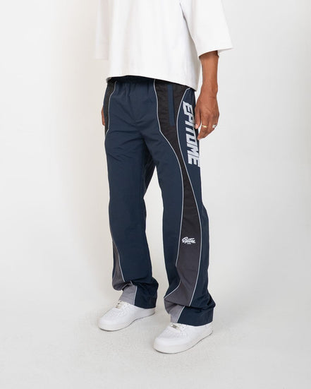 A person wearing navy and gray athletic pants with white piping and a large white vertical logo on the side, paired with a white t-shirt and white sneakers, photographed against a light gray studio background.