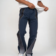 A person wearing navy track pants with dark gray side panels and white piping, paired with a white t-shirt and white sneakers, shown against a light gray studio background.