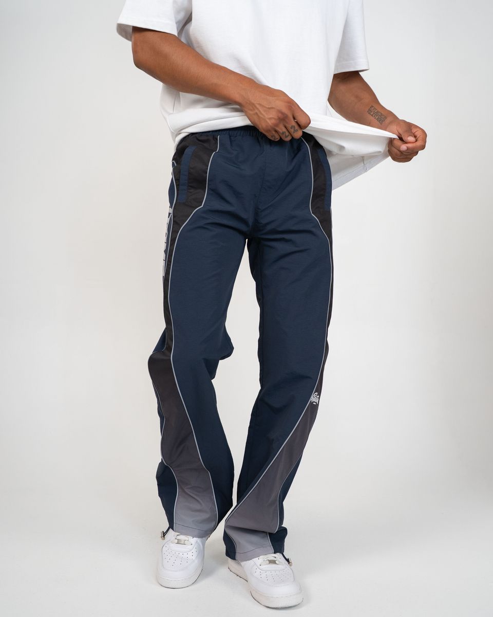 A person wearing navy track pants with dark gray side panels and white piping, paired with a white t-shirt and white sneakers, shown against a light gray studio background.