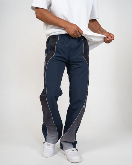 A person wearing navy track pants with dark gray side panels and white piping, paired with a white t-shirt and white sneakers, shown against a light gray studio background.