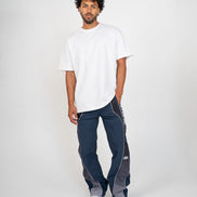 A man modeling a white crewneck t-shirt and navy blue athletic pants with gray side panels and white piping, paired with white sneakers, against a light gray studio background.
